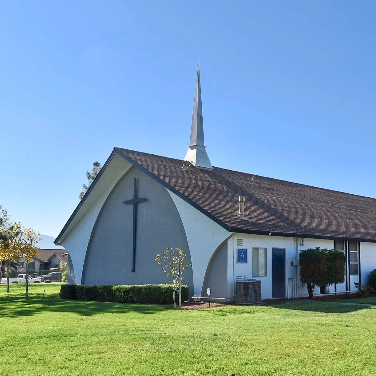 White church with a tall steeple, a cross on the front, surrounded by a grassy lawn and small trees, under a clear blue sky.