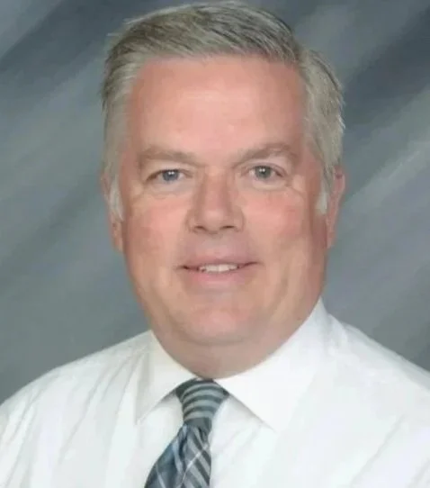 A middle-aged man with short gray hair and light skin, wearing a white dress shirt and a gray striped tie, smiling against a blue and gray gradient background.