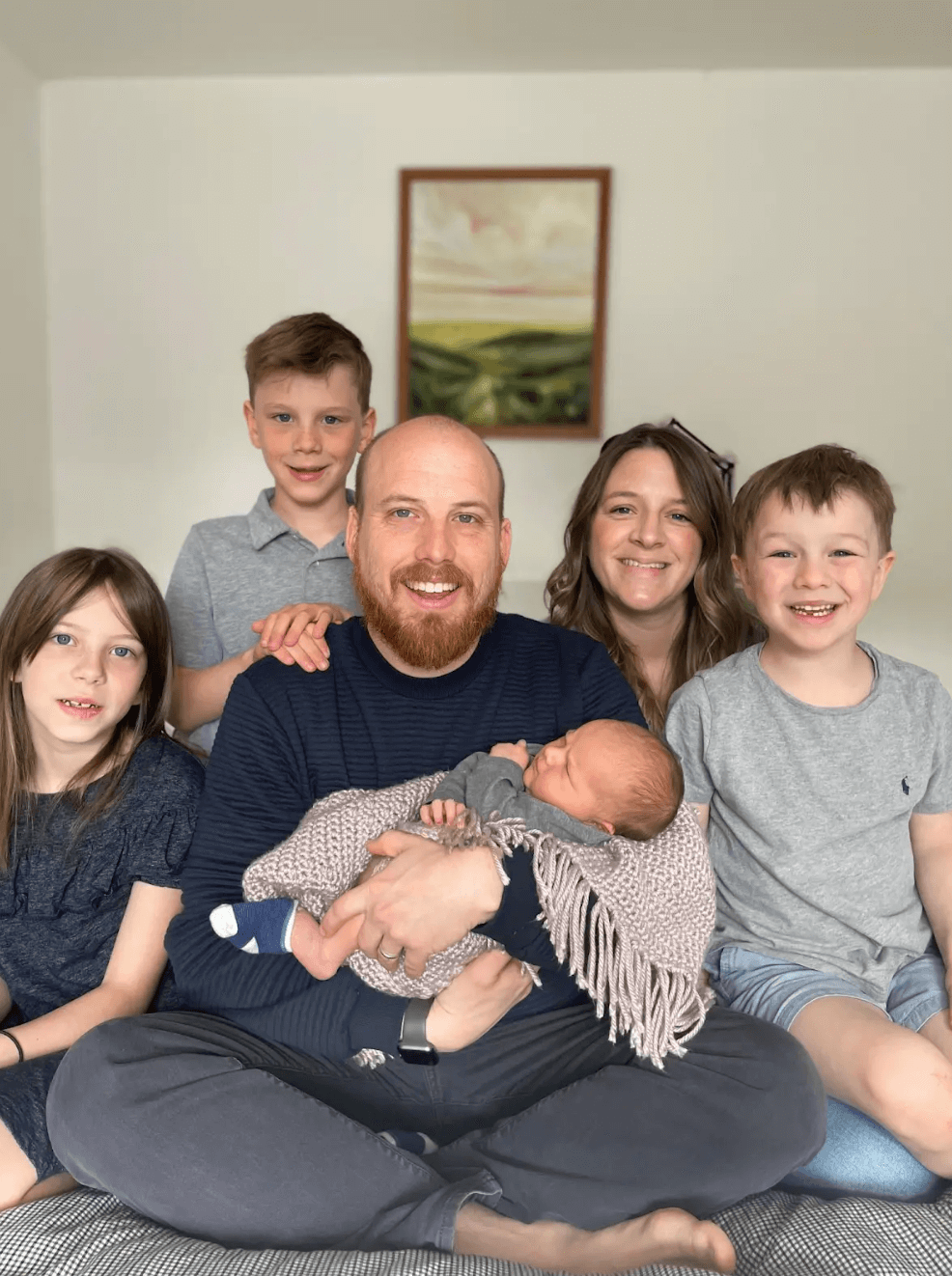 A group of seven people, including a man, woman, four children, and a newborn baby, sitting together on a couch in a living room, smiling and looking at the camera.