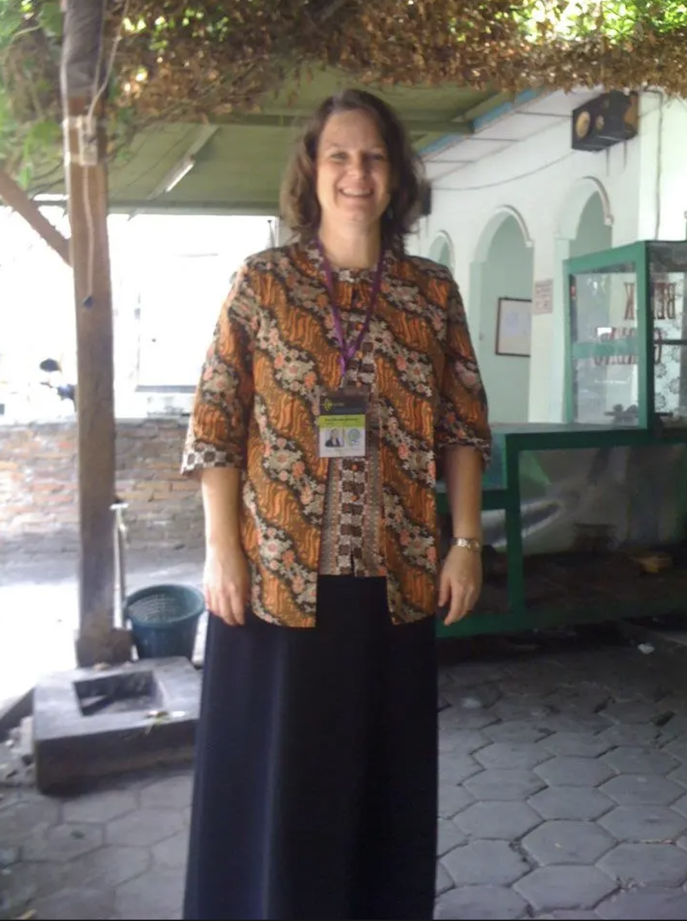 A woman with shoulder-length brown hair smiling and wearing a patterned top and long dark skirt stands indoors near a green display case.