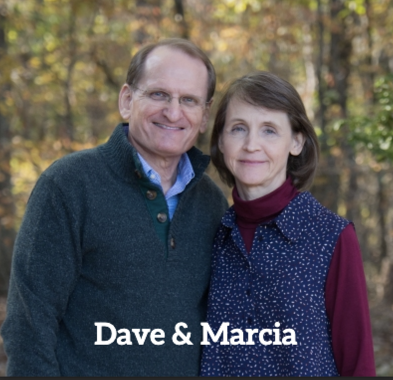 A middle-aged man and woman outdoors in fall clothing, smiling. The man wears a dark knitted sweater over a collared shirt, and the woman wears a maroon turtleneck with a blue vest. The background features autumn-colored trees. Text overlay says "Dave & Marcia."