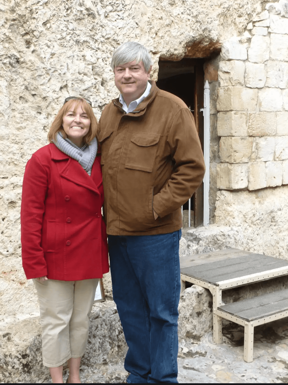 A woman and a man posing together outside a stone building. The woman wears a red coat and khaki pants, and the man wears a brown jacket and jeans.