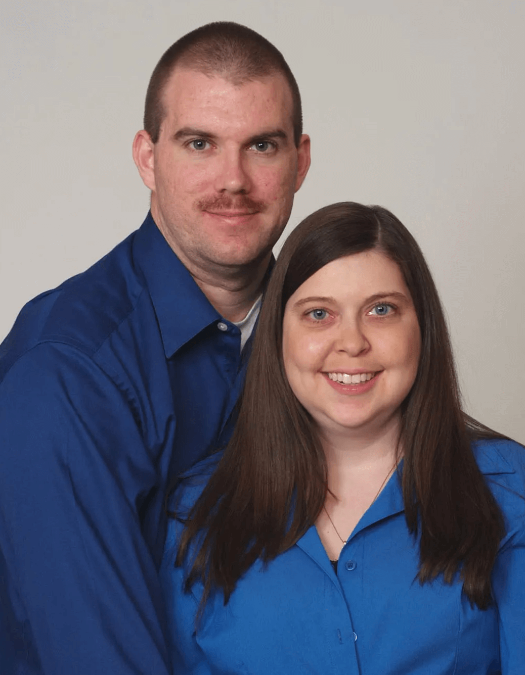A man and a woman posing together against a plain background, both wearing blue shirts, smiling at the camera.