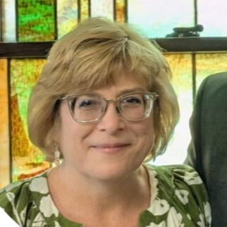 A woman with short blonde hair and glasses smiling at the camera, wearing a green and white patterned top, with a colorful window background.