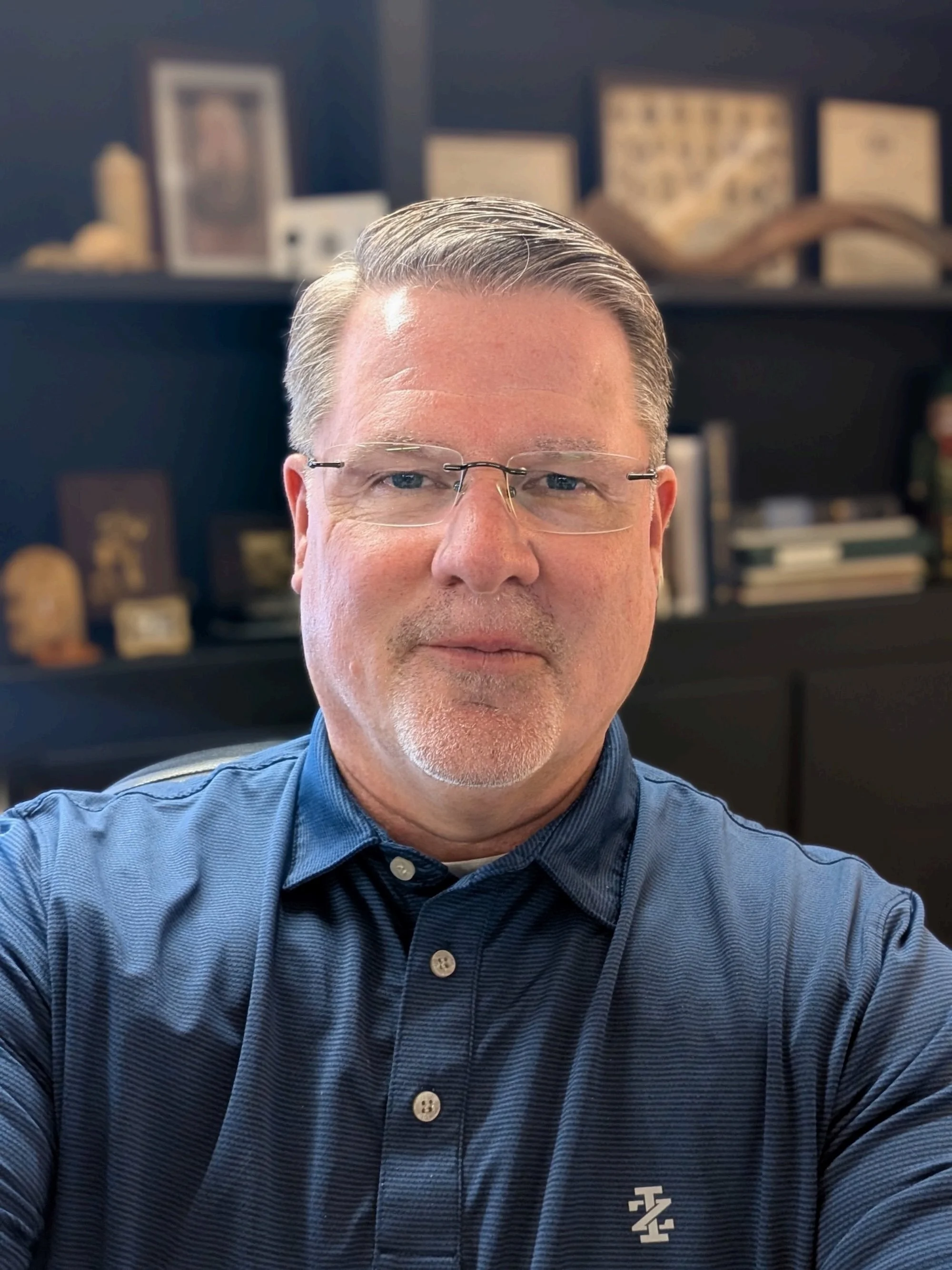 A middle-aged man with gray hair, glasses, and a blue collared shirt. Behind him is a dark shelf with framed pictures, decorations, and books.