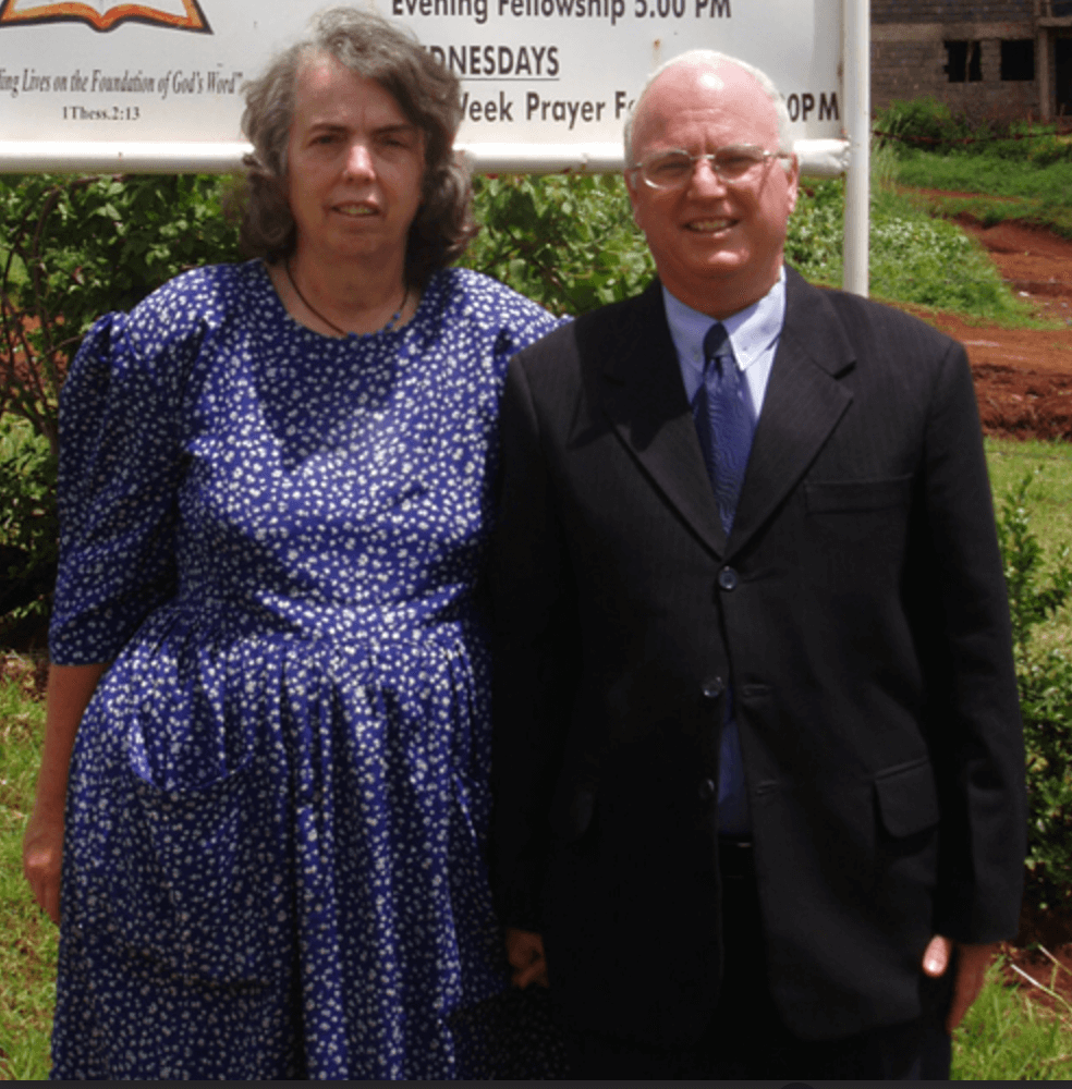 A woman in a blue dress with white floral pattern and a man in a black suit standing outdoors in front of a white sign with partial text, green foliage, and a red dirt path background.