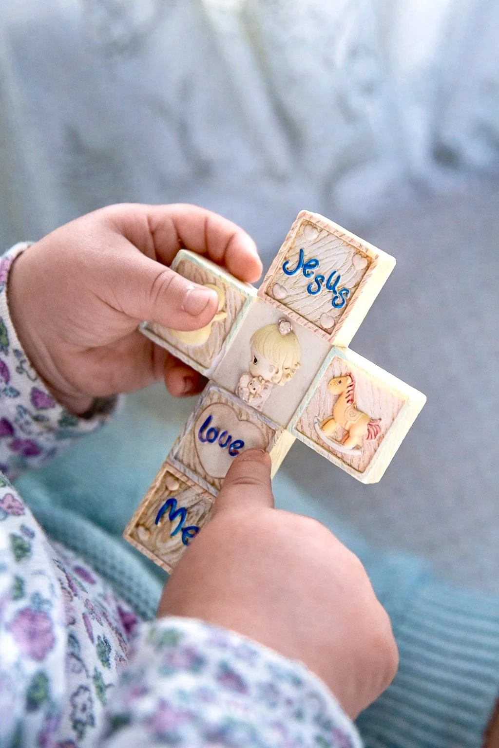 Close-up of a child's hands holding a wooden cross with small blocks. The blocks have images of a girl, a rocking horse, and words like 'Jesus' and 'love' written on them.