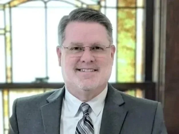 A man in a suit and tie smiling, with glasses, in front of a large window with stained glass panels.