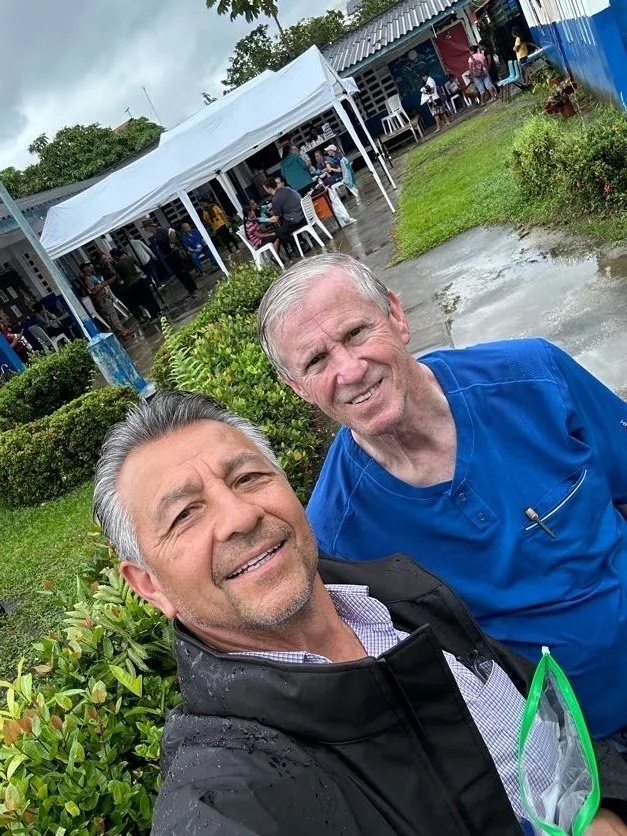 Two men smiling with an outdoor event behind them on a rainy day, surrounded by greenery and a white tent with people gathered under it.