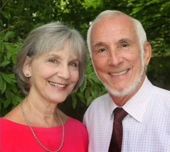 An elderly couple smiling outdoors, with green foliage in the background. The woman has short gray hair, is wearing a red top and a silver necklace. The man has short white hair and a beard, and is dressed in a white checkered shirt with a dark tie.