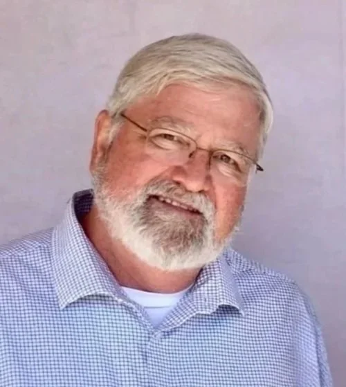 A middle-aged man with gray hair, glasses, and a beard, smiling and wearing a blue checkered shirt.