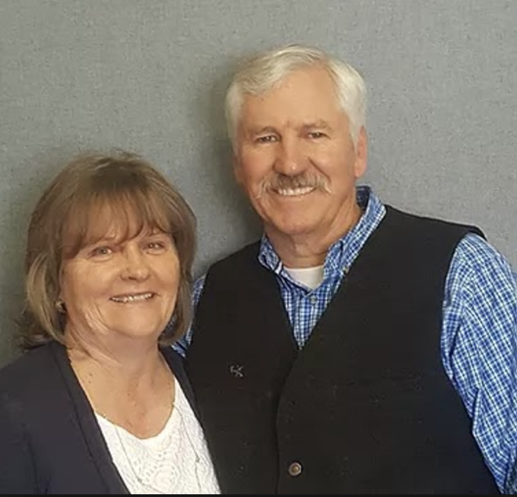 A smiling older woman with short, light brown hair and a white blouse standing next to a smiling older man with white hair and mustache, wearing a blue checkered shirt and black vest, against a plain gray wall background.