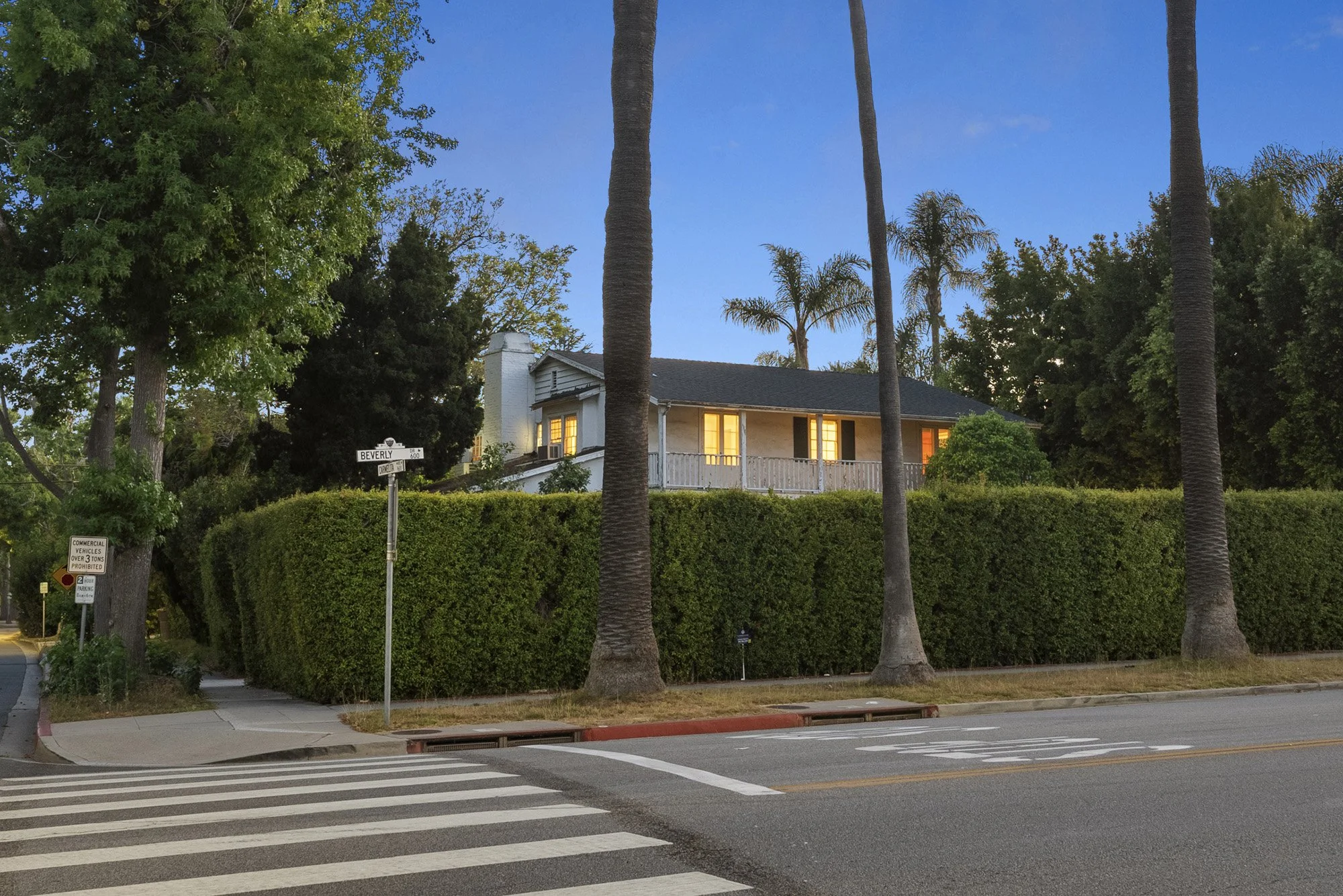 A residential street scene showing a house with a balcony and lit windows behind a tall green hedge. Several palm trees are visible, along with some other trees and bushes. A sidewalk runs along the street with a crosswalk in the foreground, and stre