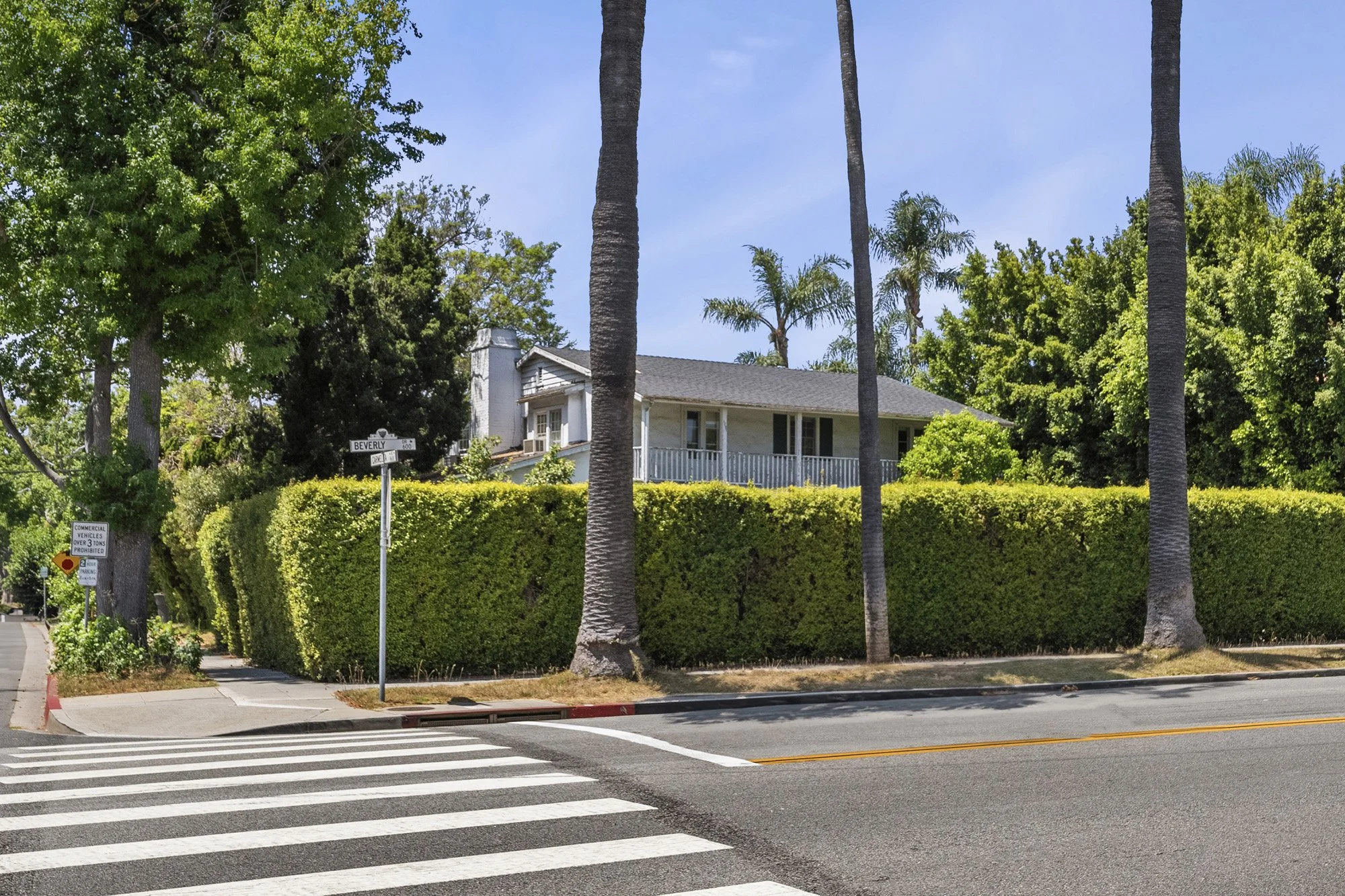 A residential street corner with a house partially visible behind a tall, green hedge. There are several palm trees and other green trees around the house. A crosswalk with white stripes is in the foreground, and there are street signs and a sidewalk
