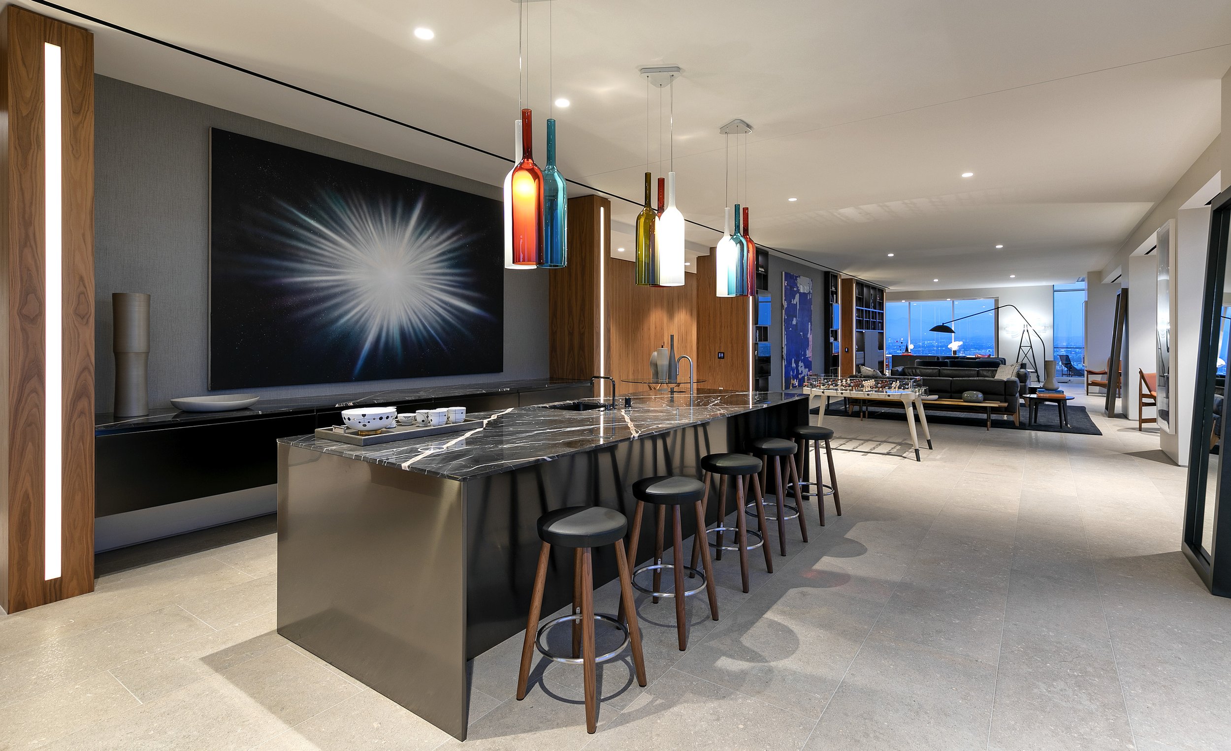 Modern open-plan living room and kitchen with a large black marble kitchen island, colorful pendant lights, a wall with a space-themed painting, and floor-to-ceiling windows showing a city skyline at night.