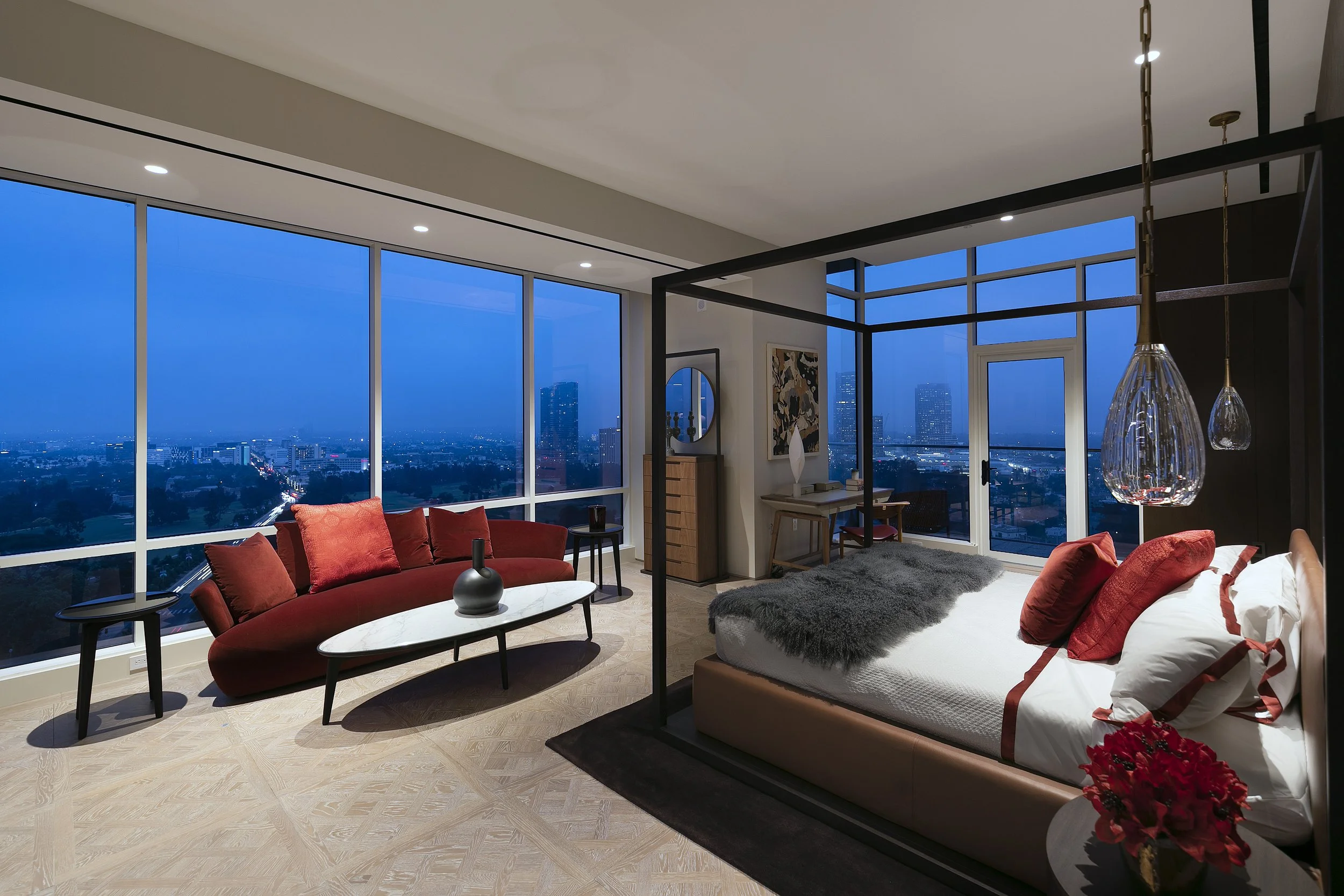Modern living room with large floor-to-ceiling windows showing a cityscape at dusk. The room has a red sectional sofa, black coffee table, and side tables, along with lamps, artwork, and a bed with gray bedding and red pillows.