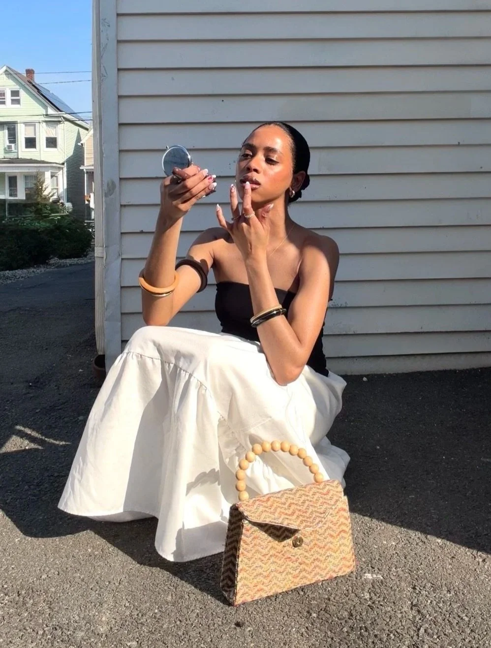 A woman sitting outdoors against a siding wall, looking into a compact mirror while applying makeup. She has a small patterned handbag with a beaded handle on the ground in front of her, and wears a black strapless top, cream-colored flowing skirt, and jewelry.