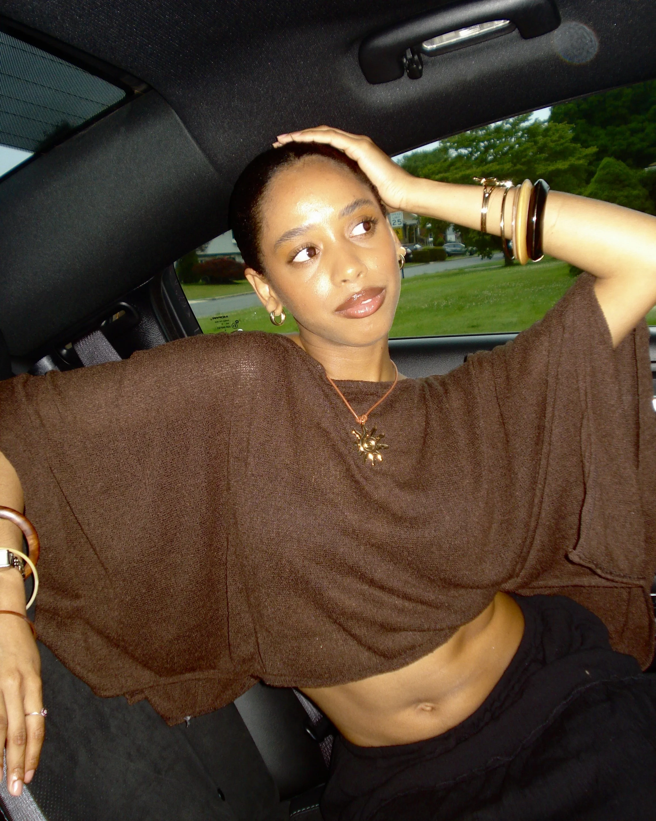 A young woman with short hair, wearing hoop earrings and several bangles, is sitting in a car with her left hand on her head and looking away from the camera. She is wearing a brown top and black shorts, and there is greenery visible outside the car window.