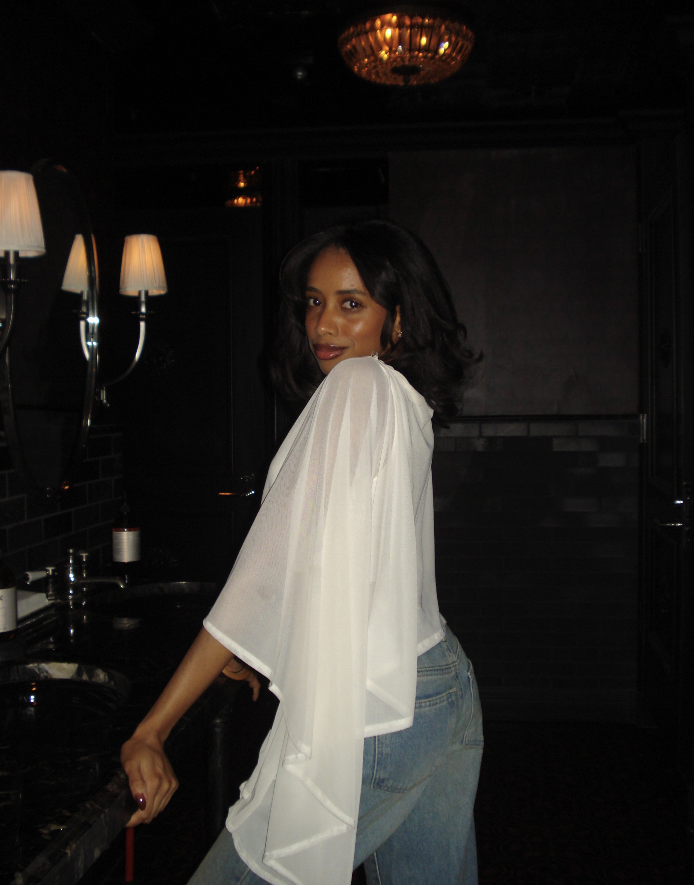 A woman with shoulder-length black hair and medium skin tone wearing a white shirt and light blue jeans, leaning on a black countertop in a dimly lit room with black walls, a chandelier hanging from the ceiling, a lamp with a white shade, and a mirror reflecting part of the room.