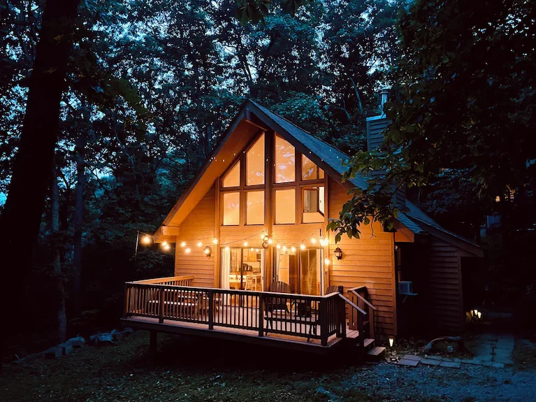 A cozy A-frame cabin with large front windows, surrounded by trees, with a warmly illuminated deck and string lights at dusk.