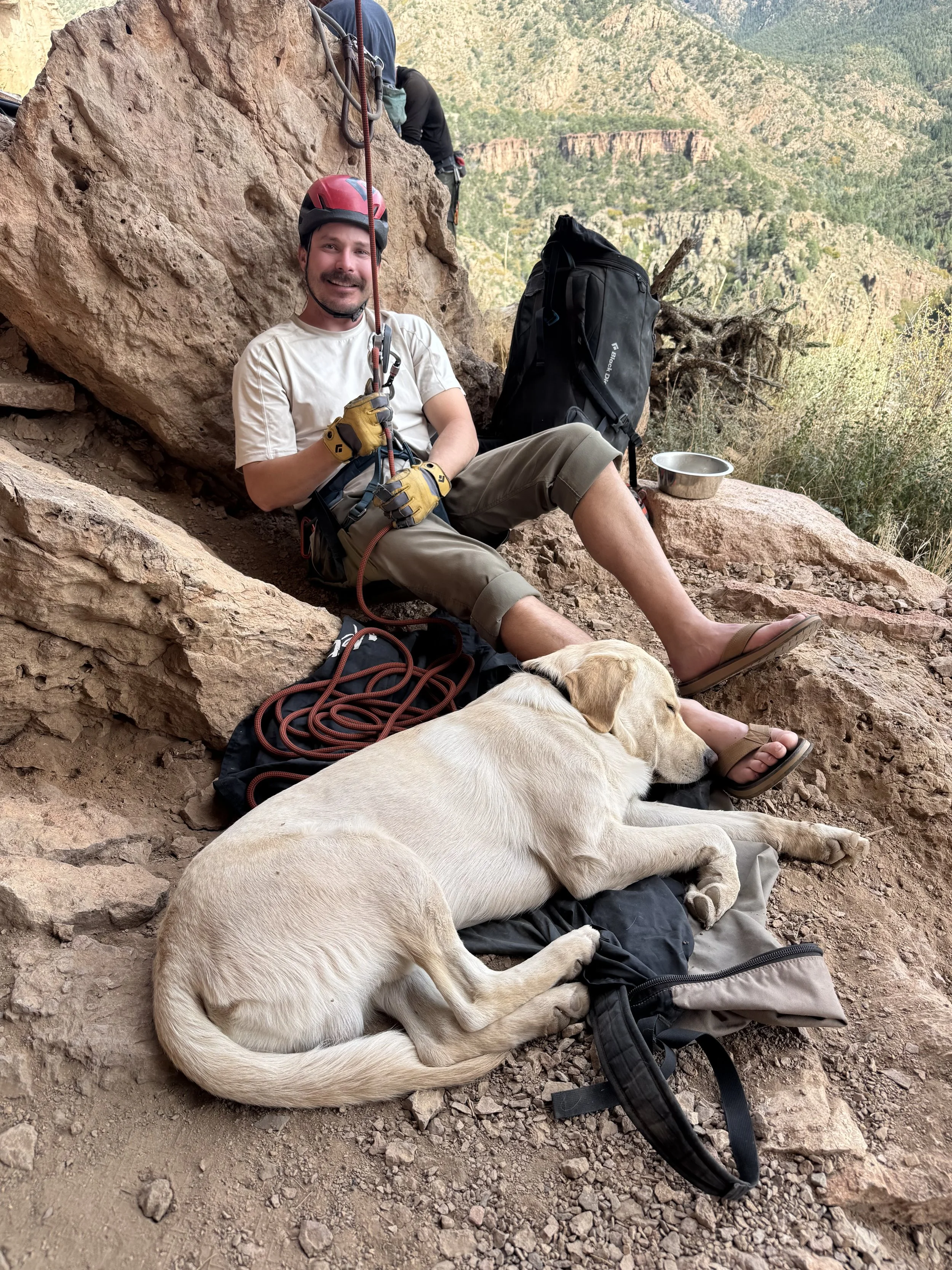 A man in a red helmet and tan shorts sitting on rocky ground beside a yellow Labrador Retriever dog that is resting. The man is smiling, appears to be on a hiking or climbing trip with various outdoor gear and a black backpack nearby. The background shows a mountainous landscape with green vegetation.