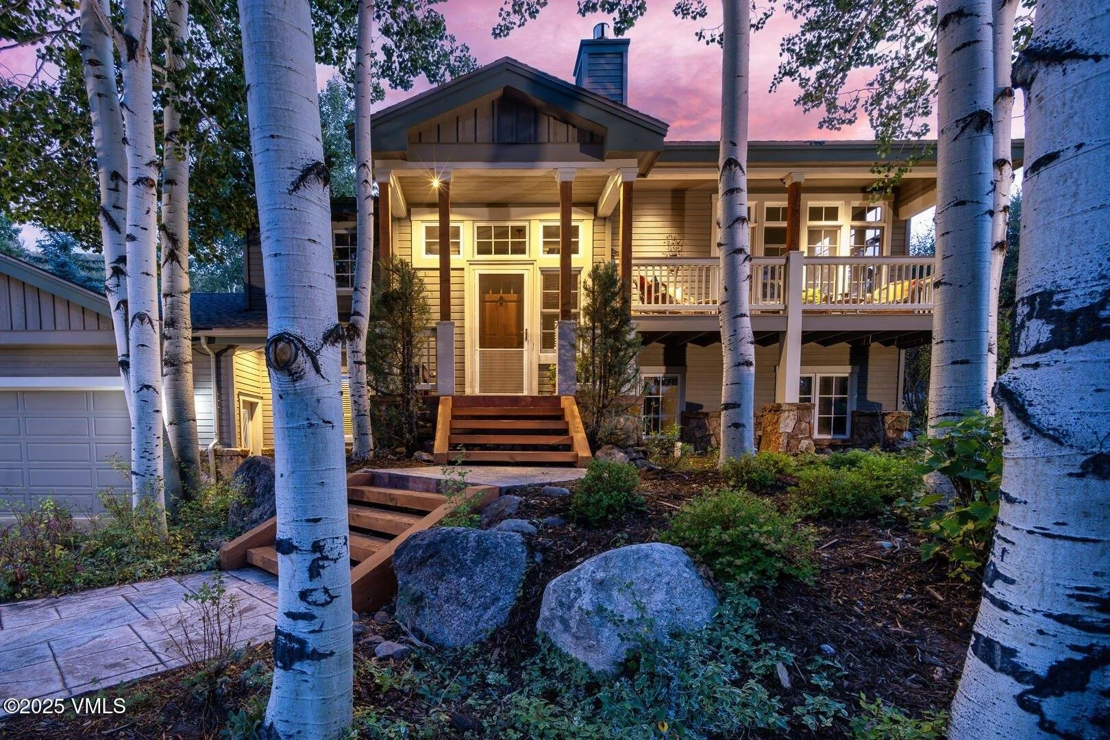 A modern house with a large wooden front porch and rock landscaping, surrounded by tall white birch trees, during twilight.