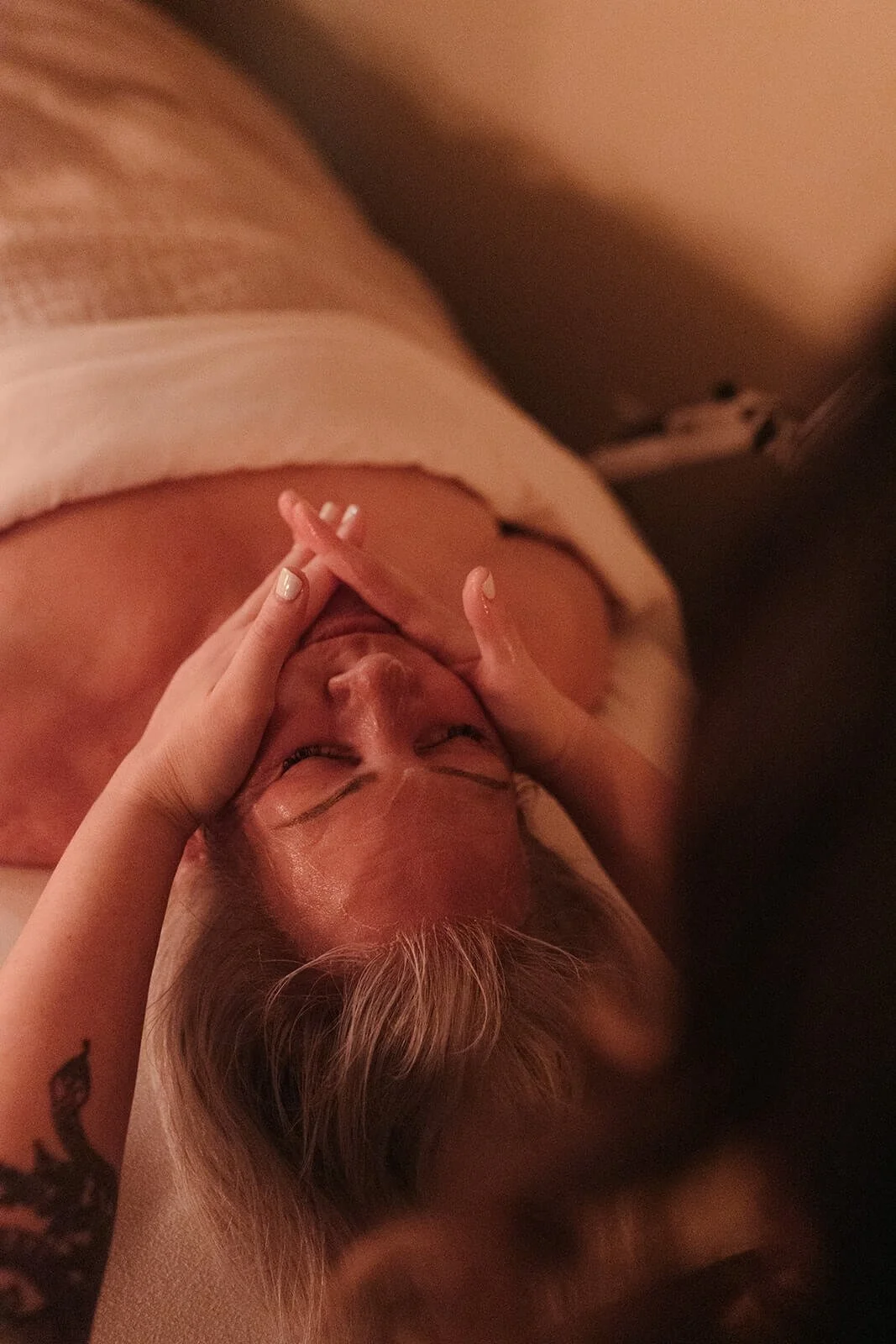 A person lying down, eyes closed, receiving a facial massage, in a dimly lit room, with a tattoo visible on their left arm.