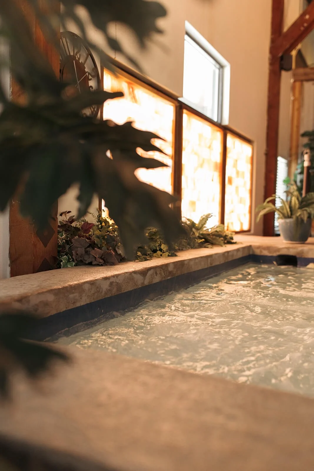 Indoor hot tub with bubbling water, decorated with green plants and ivy, illuminated by warm Himalayan salt panel in the background.