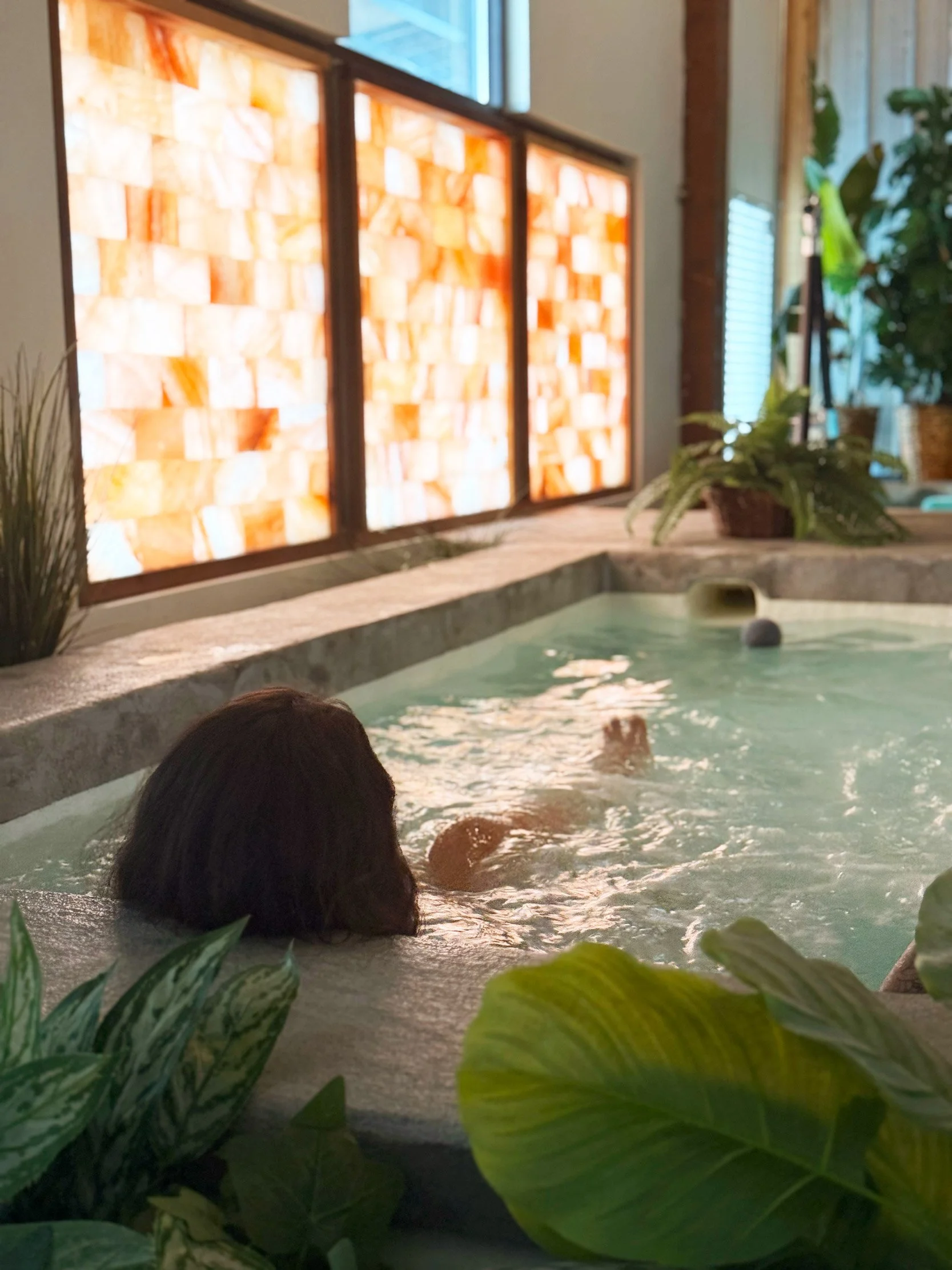 A person with dark hair lounging in a hot tub or pool indoors, surrounded by green plants. There is a Himalayan salt panel glow in the background.