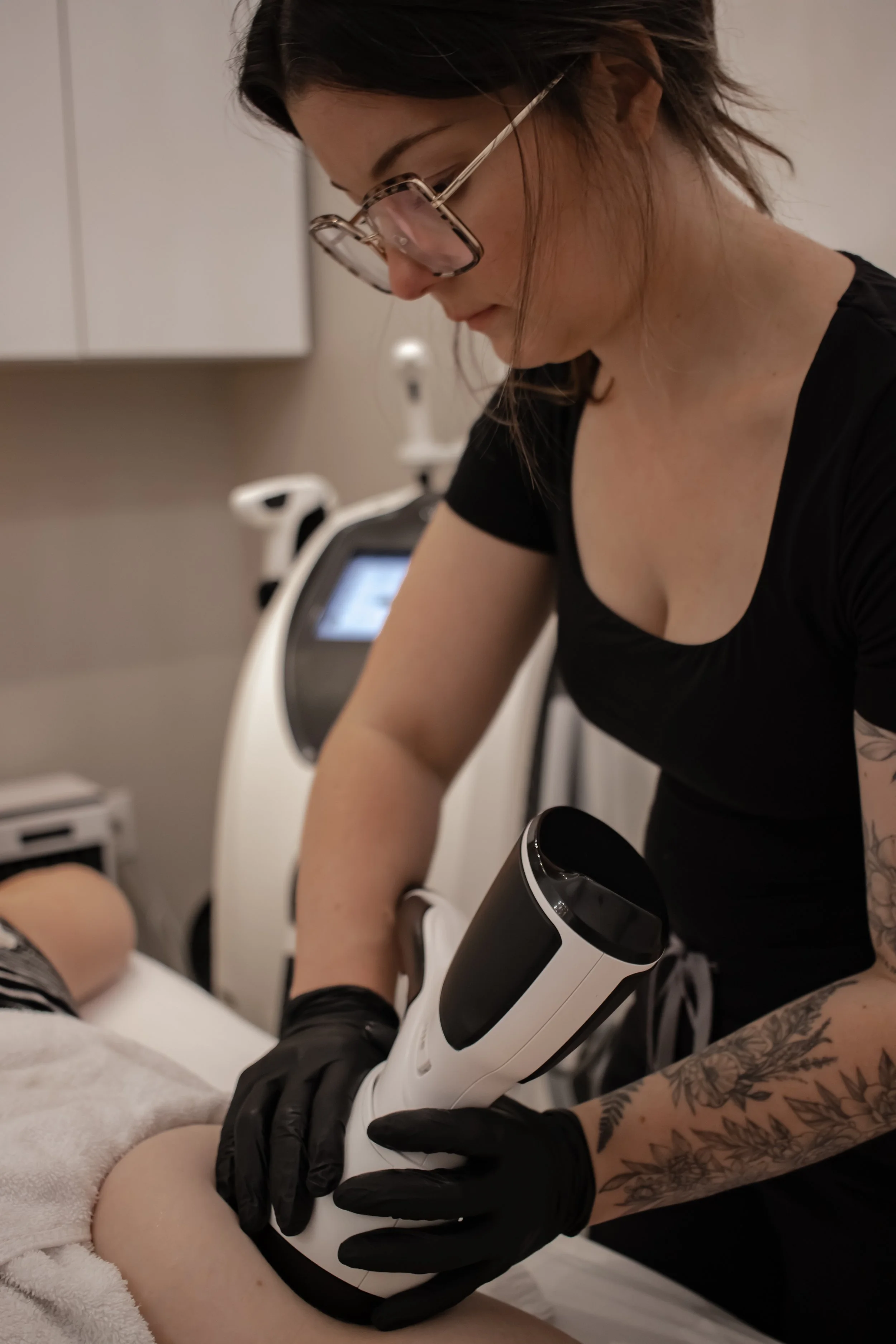 A woman with glasses and tattoos on her arm performing a cosmetic procedure using a handheld device on a person's leg, in a clinical setting.