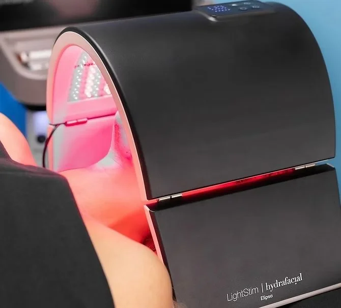 Person receiving a light therapy facial treatment with a specialized device emitting pinkish light.