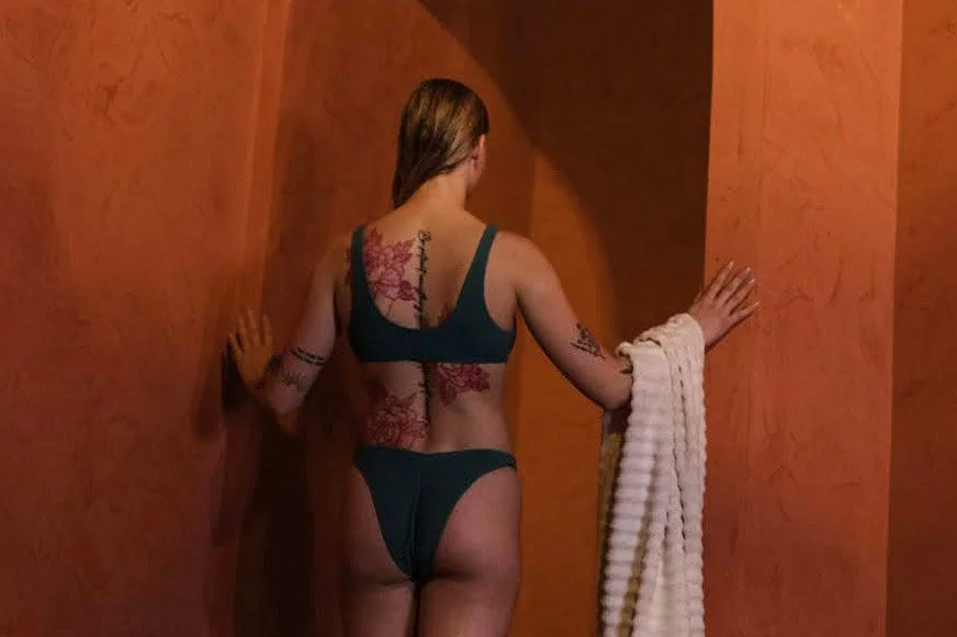 Back of a woman with tattoos, wearing a dark green bikini, holding a white towel, entering a spa shower stall.