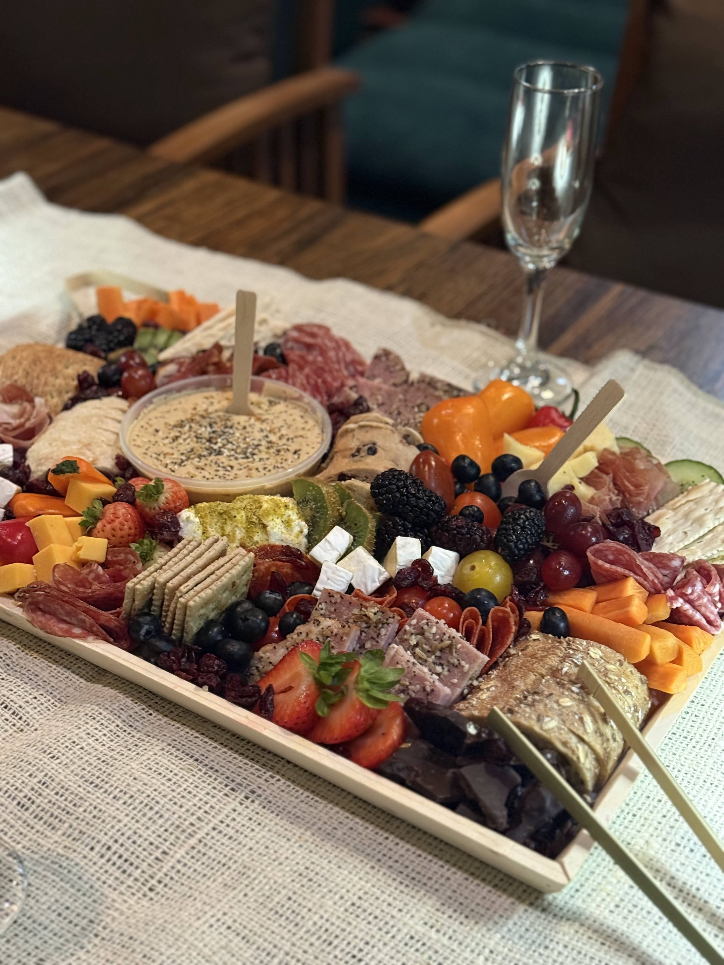 A large charcuterie board filled with various cheeses, fruits, vegetables, meats, and dips, with a champagne glass nearby on a wooden table.