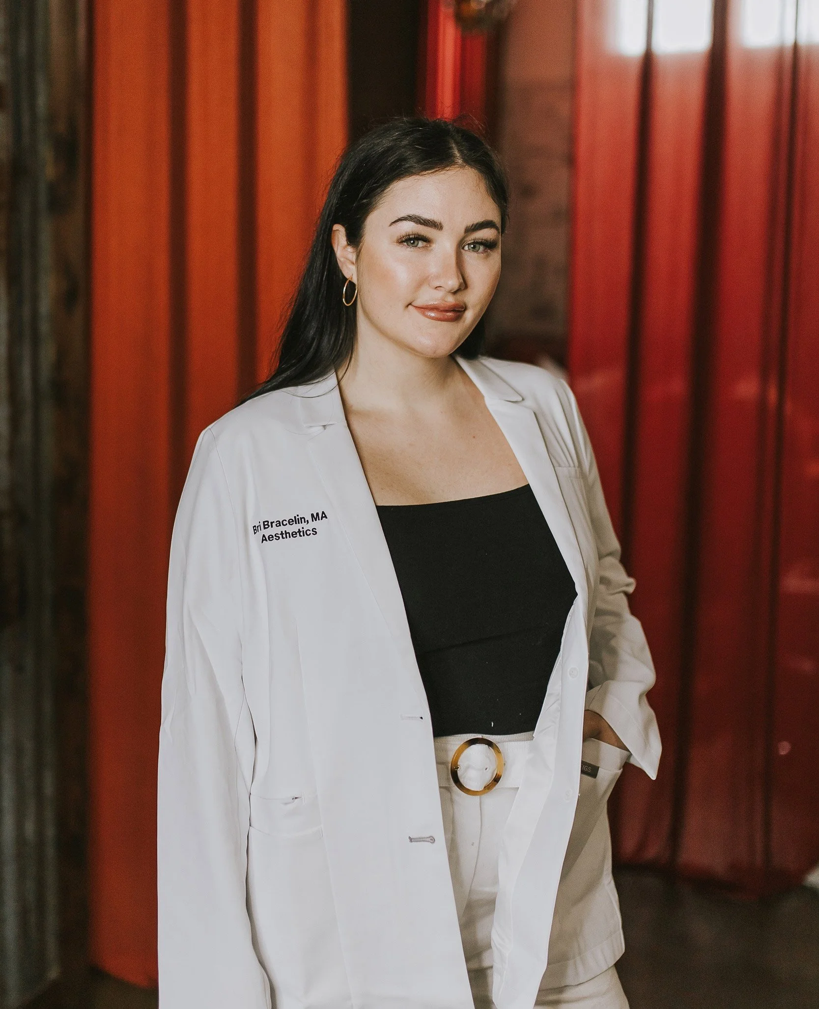 Spa owner, Bri Bracelin, in a white lab coat standing in front of red wooden wall panels, smiling and looking at the camera.
