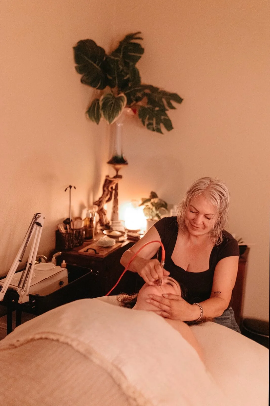 A woman receiving a facial treatment from a therapist in a cozy room with warm lighting, plants, and decor.