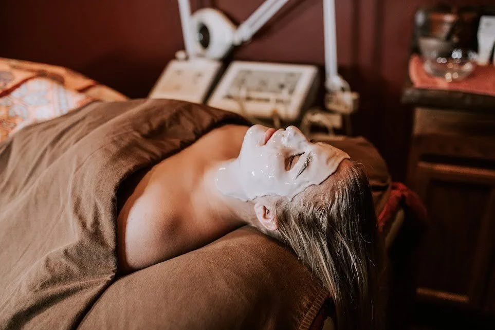 Woman lying on a bed with her eyes closed, receiving a facial treatment with a mask applied to her face.