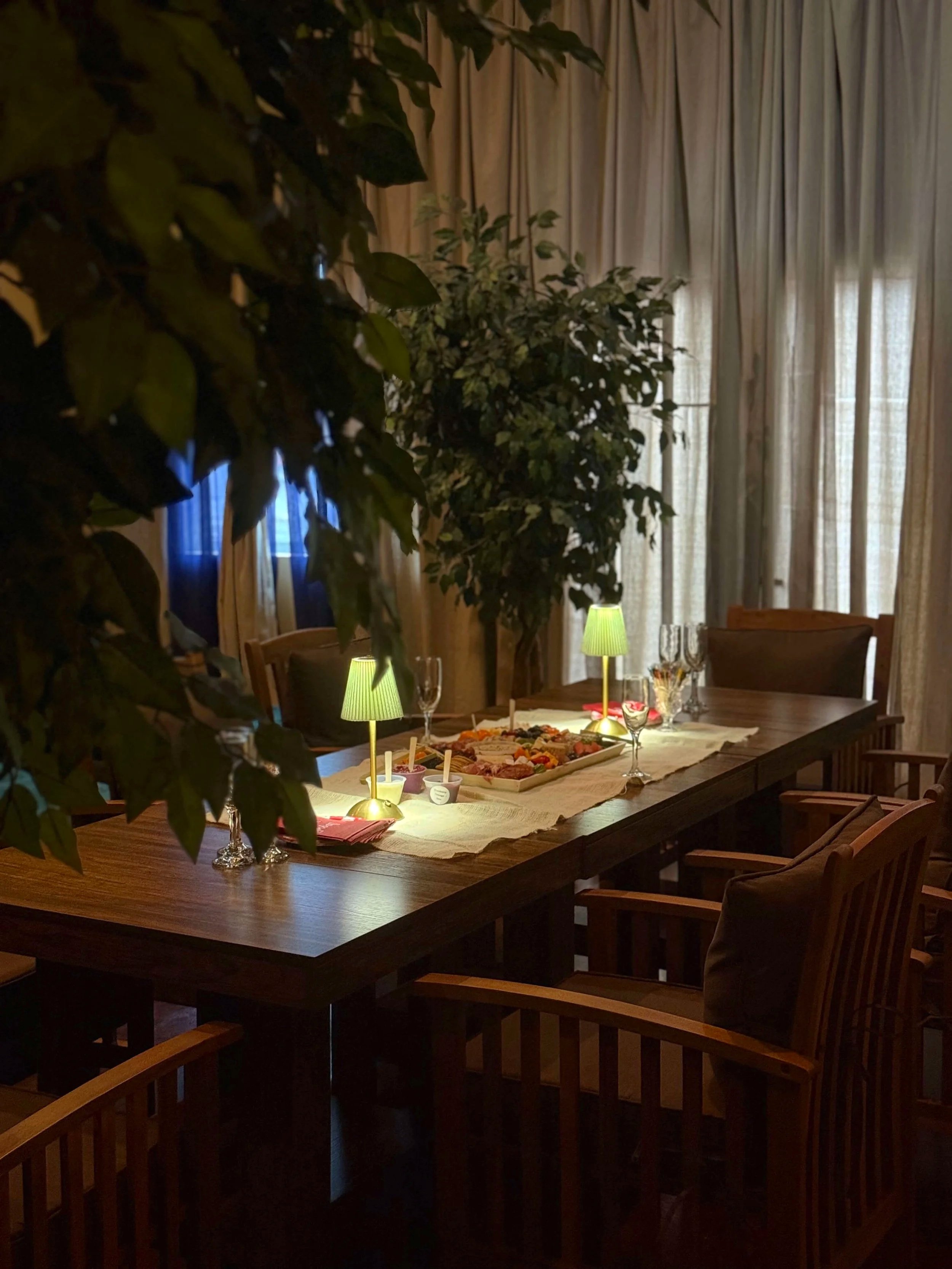 A dining room with a large wooden table, surrounded by wooden chairs, decorated with a buffet of appetizers and small dishes, illuminated by two small table lamps. Tall beige curtains cover the windows.