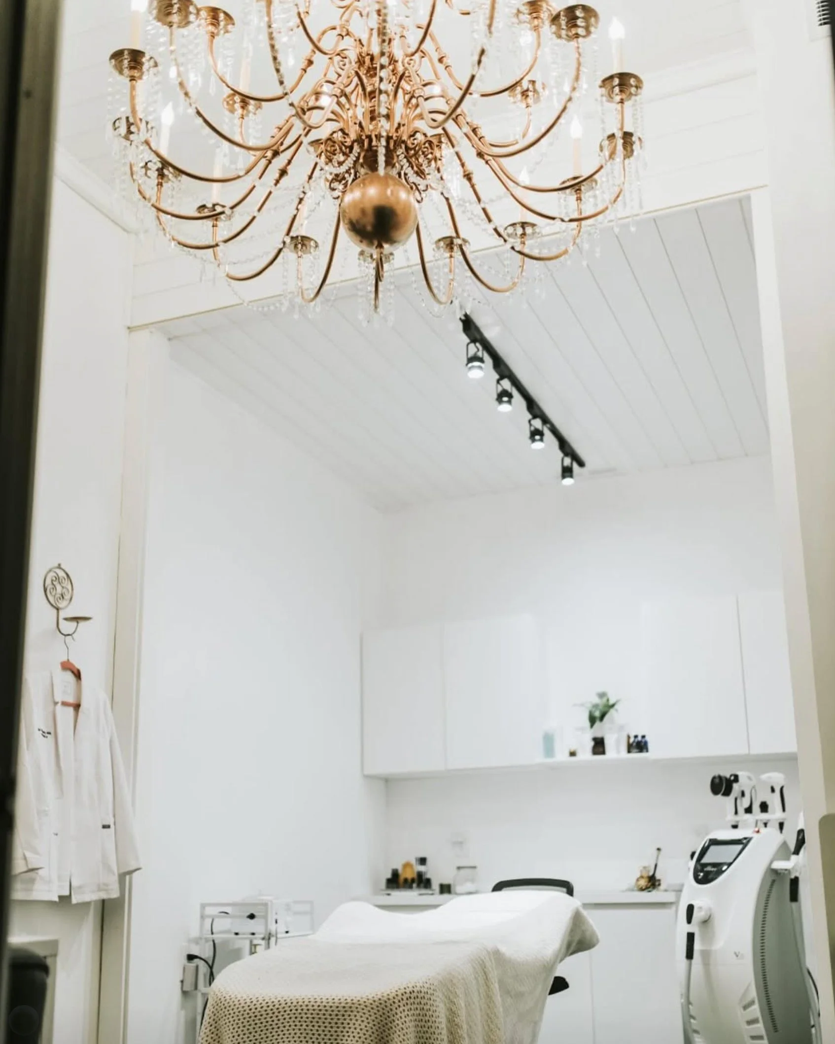 Medical or dental room with white walls, a large ornate chandelier hanging from the ceiling, a bed covered with a cream-colored blanket, medical equipment including a chair, sterilizer, towels, and various tools, as well as medical devices lined up on the counter and a cabinet with shelves.
