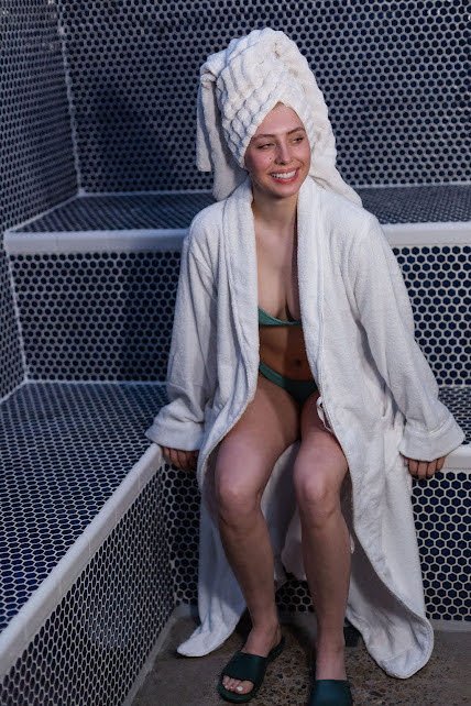 A woman sitting in a steam room with her hair wrapped in a towel and wearing a robe, towel, and slippers.