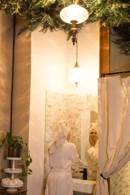 Woman in a bathroom with a towel on her head, looking into a mirror. Decor includes hanging lights, greenery overhead, a curtain, and a plant on a small table.
