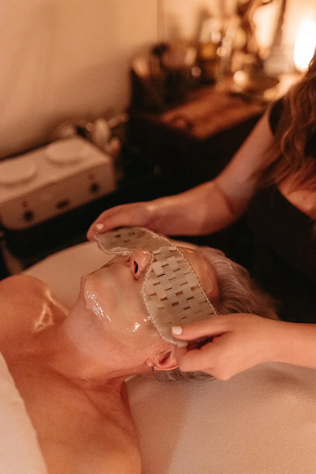 A woman is receiving a facial treatment, with a sheet mask over her face, in a dimly lit room.