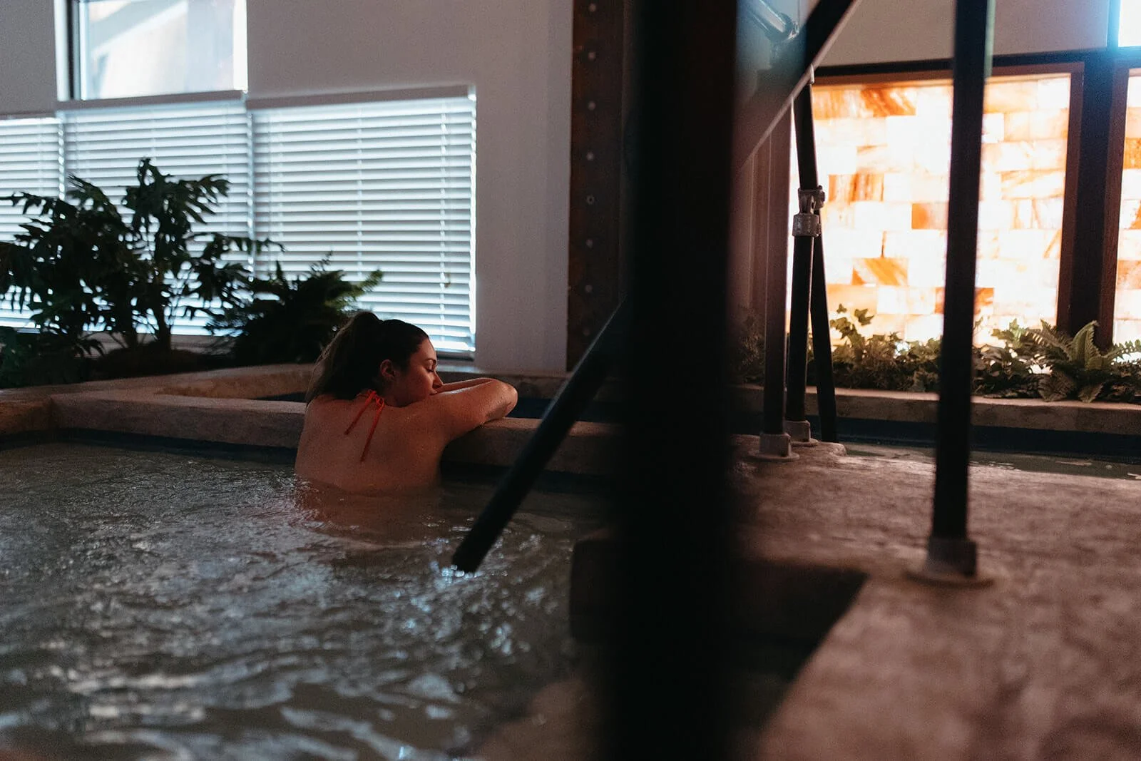 A woman relaxing in a hot tub indoors, leaning on the edge with her head resting on her arms, surrounded by plants and window blinds with natural light coming in.