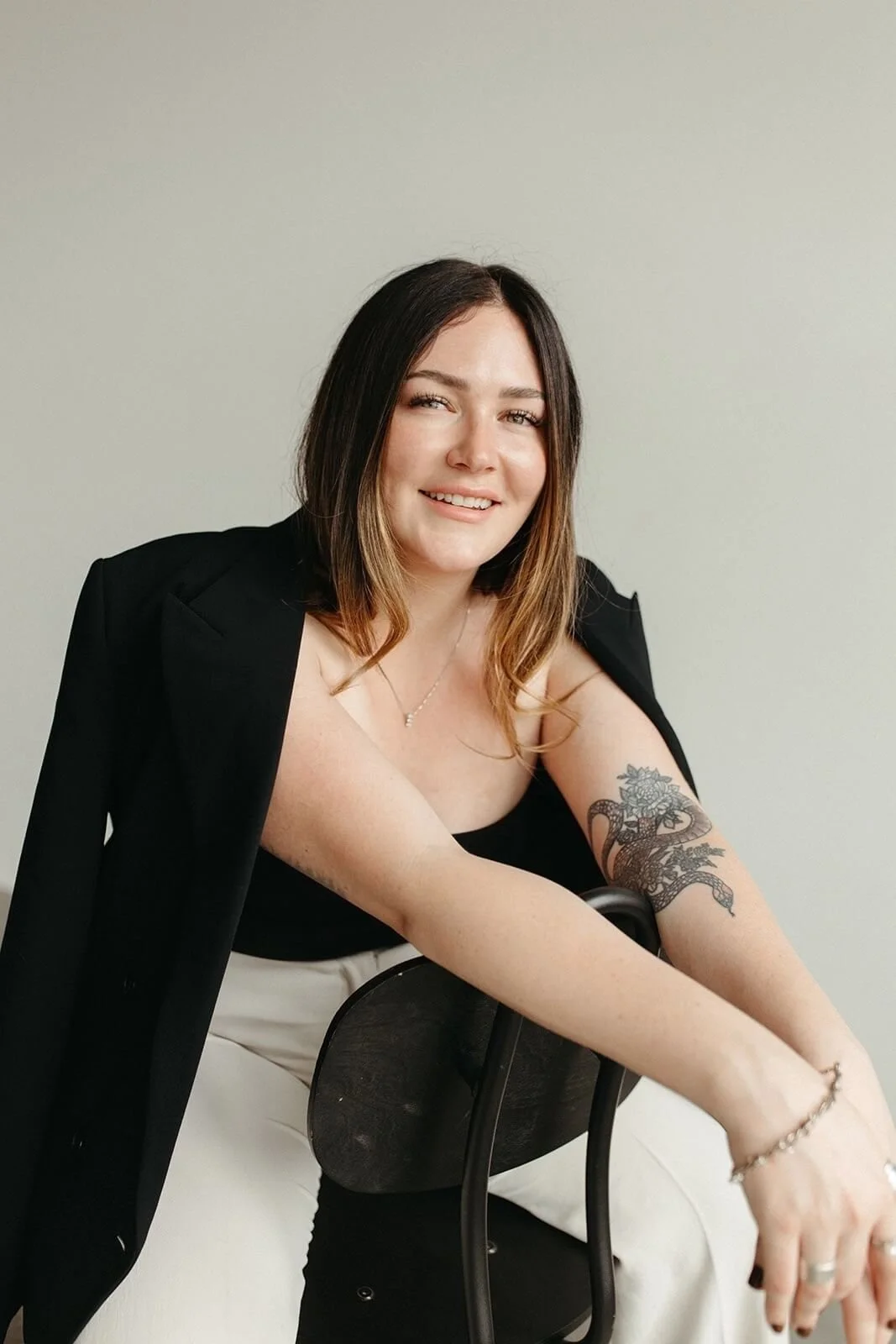 Spa Bliss owner, Bri Bracelin, wearing a black sleeveless top, a black jacket draped over her shoulders, beige pants, jewelry, and a tattoo of a snake and flowers on her right arm, sitting on a black chair against a plain wall.