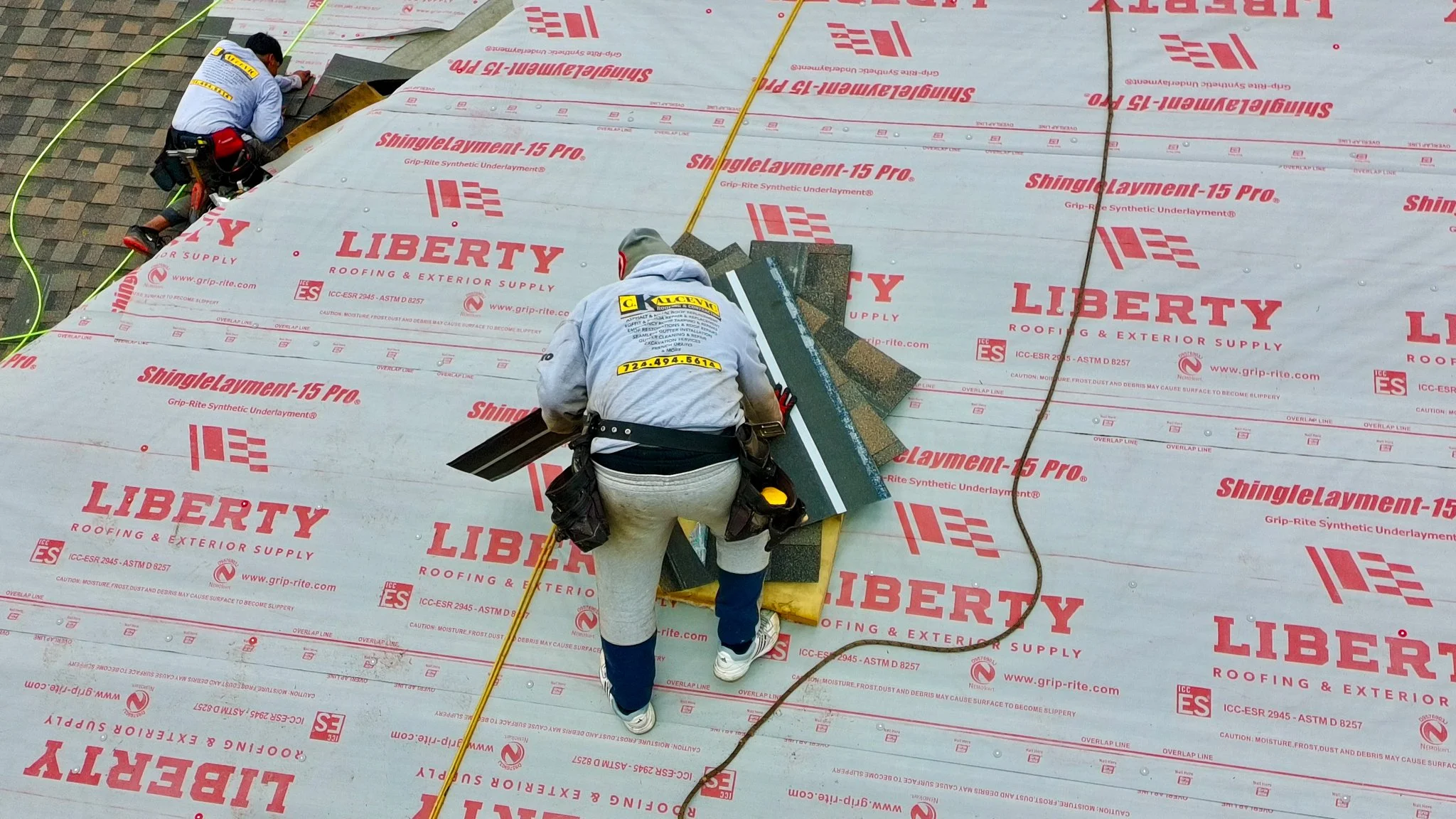 Two Pittsburgh construction workers installing roofing material on a building roof. They are working on a surface covered with Liberty brand roofing underlayment, with one worker in focus laying shingles over the underlayment. The workers are wearing