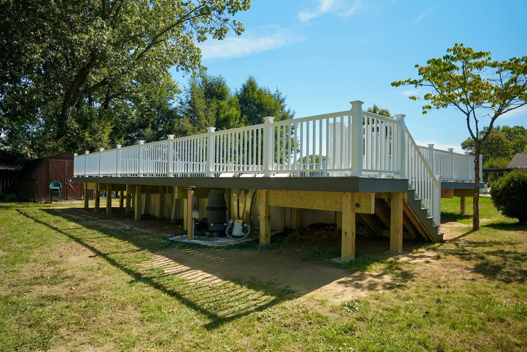 A newly built raised deck with white railing, outdoor stairs, and wooden support beams in a backyard with green grass and trees.