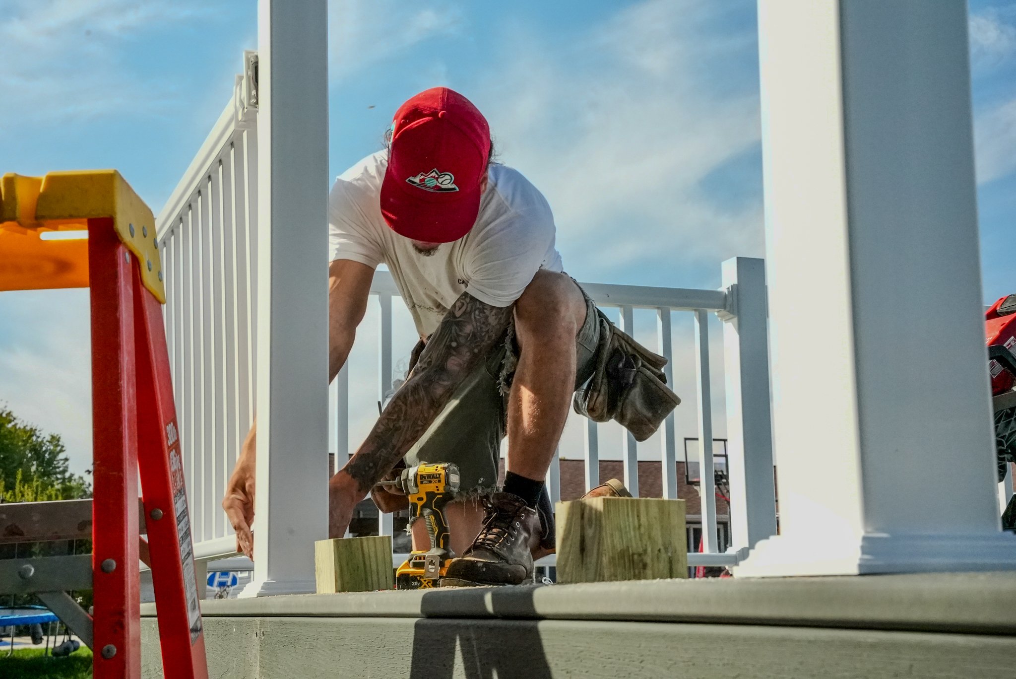 A man in a red cap, white t-shirt, and shorts working on a construction project on a porch, using a drill and wooden blocks, during daytime.