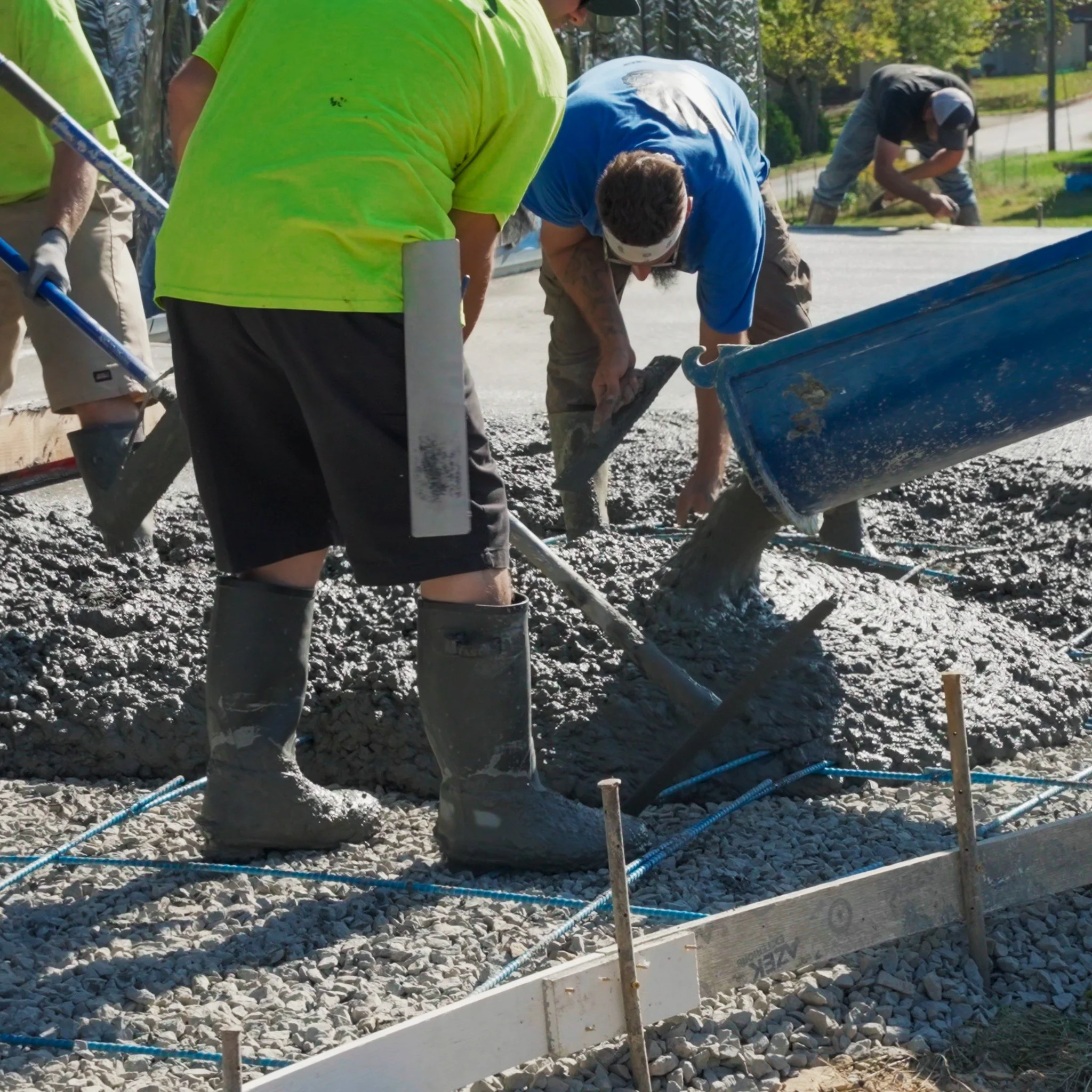 Pittsburgh contractors pouring and spreading concrete on a construction site, with some using shovels and a concrete chute, wearing work clothes, boots, and gloves.