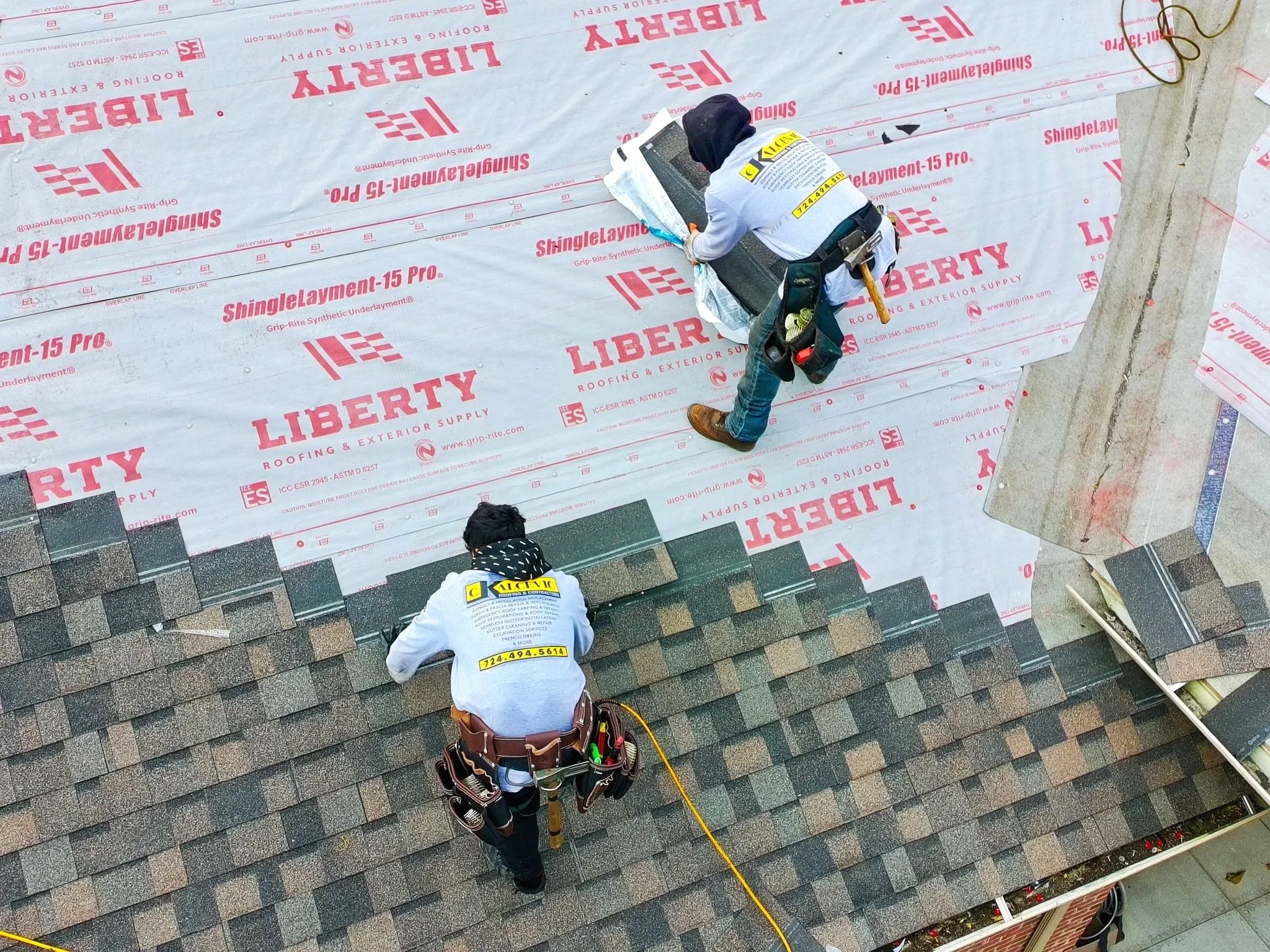 Two Pittsburgh roofers installing roofing shingles on a roof, with a layer of underlayment marked 'Liberty' and one worker placing shingles, both wearing safety harnesses and tools.