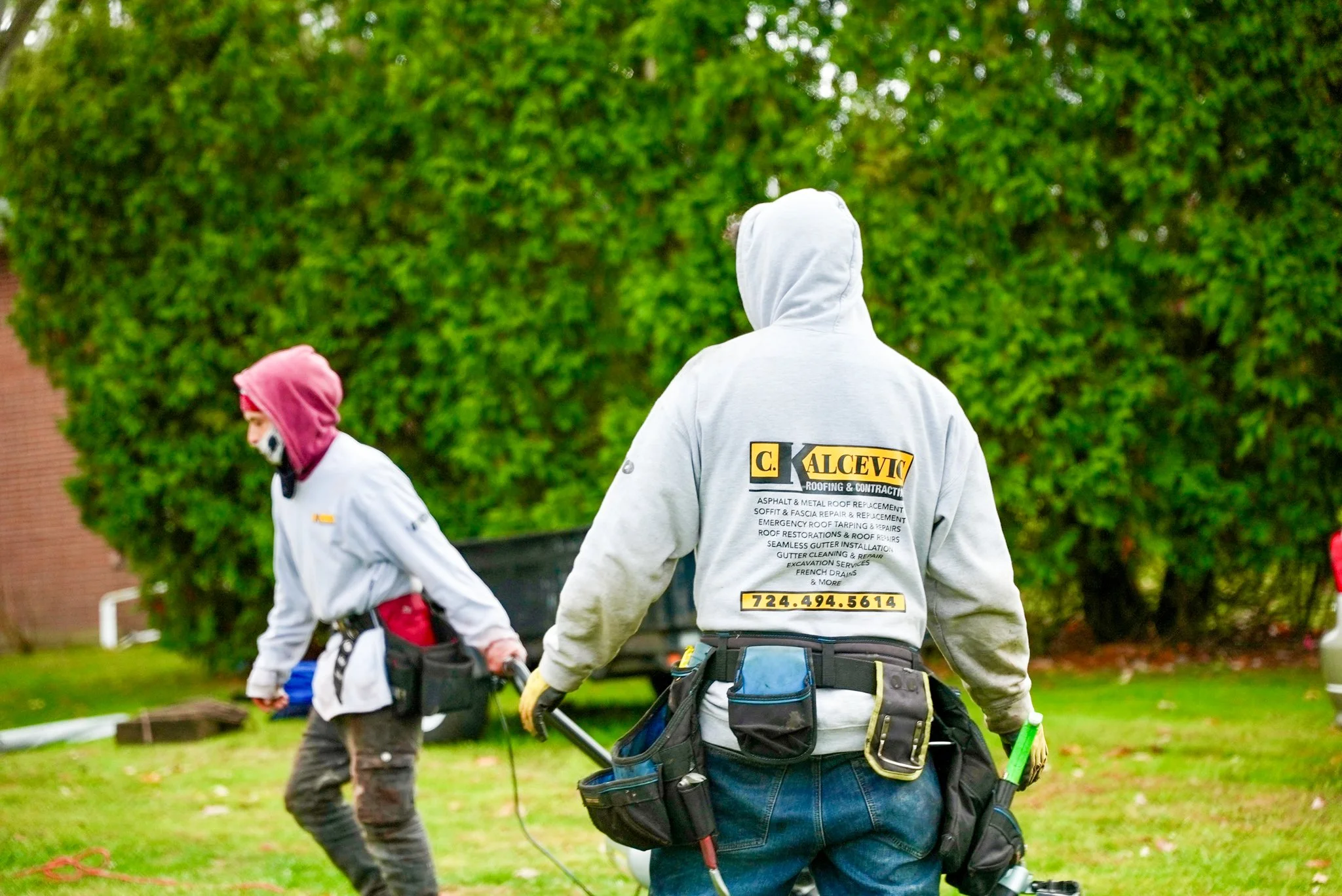 Two Pittsburgh roofers working outdoors, one with a white hoodie with a business logo and the other with a red hoodie, working on a roof repair project.