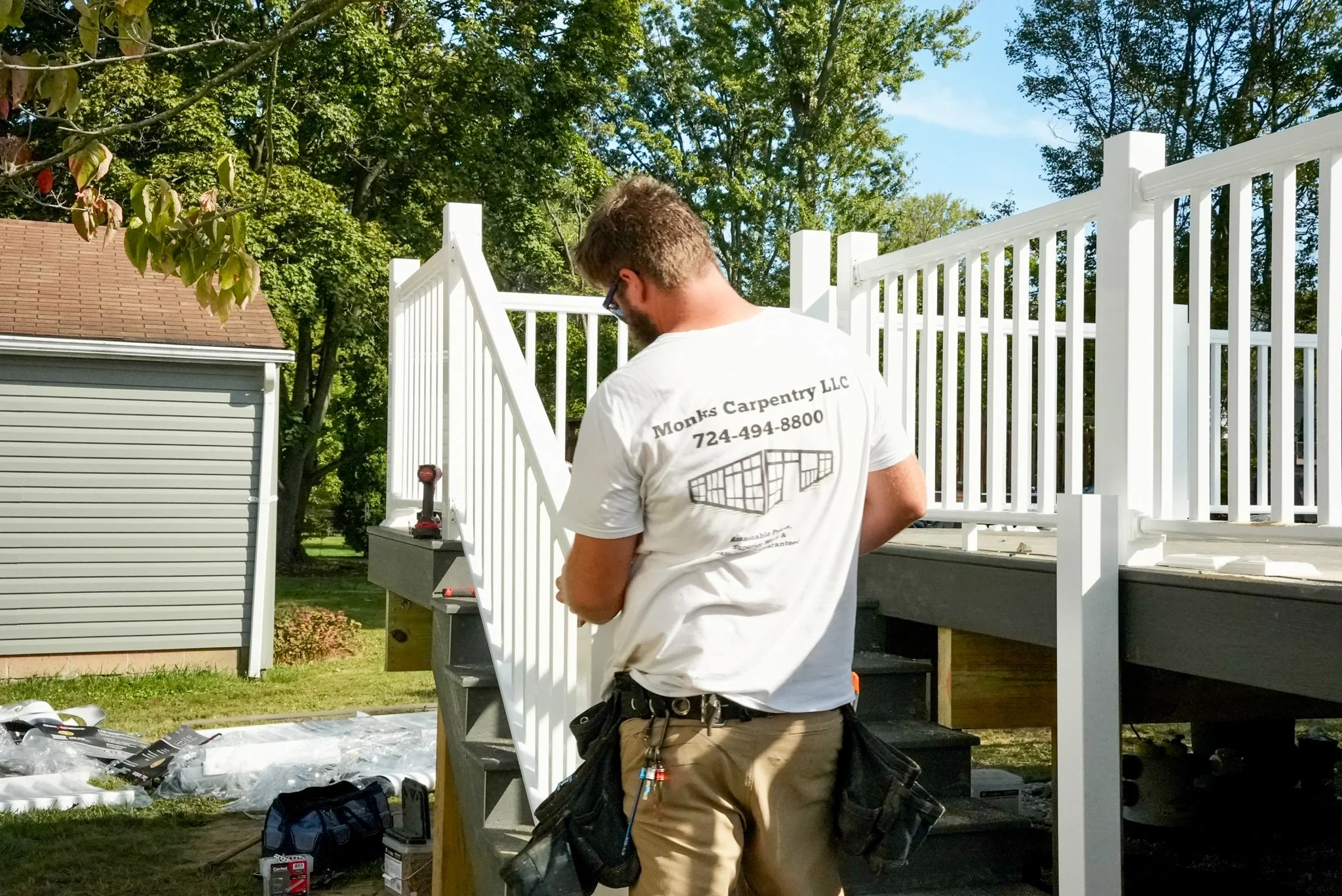 A construction worker with a white t-shirt and tan pants standing next to a white deck railing, working on building or installing a deck, with tools and materials nearby on the grass.